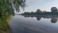 Morning on the river. A light mist and rays of sunshine. Tall grass and trees grow along the banks and are reflected in the calm w