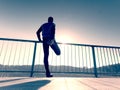 Morning runner in tall black leggings makes body stretching on bridge path. Outdoor exercising