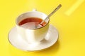 Morning sun shines to white porcelain cup with hot black tea, just brewed, silver spoon in, on yellow board