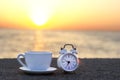 Morning sunrise on the beach. White cup and alarm clock on stone breakwater with sea background and sunshine. Space for text.