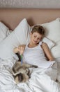 Cute handsome blonde boy lying in white bed. Boy smiling and playing with cat. Relax time Royalty Free Stock Photo