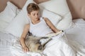 Cute handsome blonde boy lying in white bed and reading a book. Boy smiling and playing with cat. Relax time Royalty Free Stock Photo