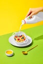 Morning vibe. Female hand pouring milk into bowl with sweet muesli, cereal with berries against green yellow background Royalty Free Stock Photo