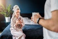 Woman in bathrobe, towel on her head sits on bed, using smartphone. In foreground is man with cup of coffee in his hands Royalty Free Stock Photo