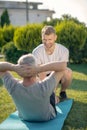 Smiling brown-haired male helping grey-haired male with sit ups