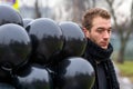 Birthday is a sad holiday concept. A young blonde man wearing black scarf and coat holding black balloons going down street Royalty Free Stock Photo