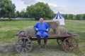 Cheerful man in medieval blue clothes is sitting on a horse-drawn cart on the background of a green