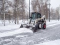 A small loader with a bucket removes snow from a sidewalk in a city park