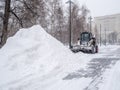 A small loader with a bucket removes snow from a sidewalk in a city park