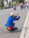 A gray-haired elderly Asian man squats down, taking pictures on his phone