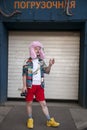 A young man in a motley Hawaiian-style shirt, white T-shirt, red shorts and a pink fur hat with a camera stands in front of the