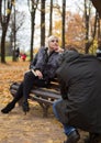 A man shooting young attractive woman sitting on the bench in autumn park. Royalty Free Stock Photo