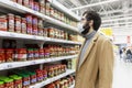 Moscow, Russia, 07/17/2020: A masked man in a supermarket dressed in canned food. Young brunette with a beard in a beige coat.