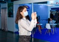 A dark haired woman in a protective medical mask stands indoor with smartphone in her hands