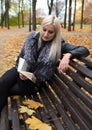 A young attractive woman wearing fur black jacket holding book sitting on the bench in falling park. Royalty Free Stock Photo