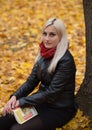 A young attractive woman wearing fur black jacket holding book sitting by the tree. Walking around autumn park. Royalty Free Stock Photo