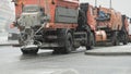 Winter, it snowing. On road, two orange cars are driving to clean up the roads