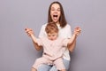 Happy woman with toddler child sitting sitting together isolated over gray background, mommy holding kid's hands