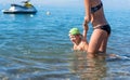 Mother in black sunglasses and smiling baby boy son in green baseball cap playing in the sea in the day time. Positive human emot Royalty Free Stock Photo