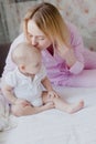 Mother blonde in pink pajamas kisses her infant sitting on her bed