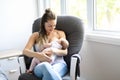 Mother is breastfeeding a baby sitting on a chair close to the window