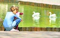 Mother and child relaxing in the park by the pond. They sitting on a sidewalk by the lake and looking on water with two white swan