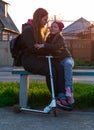 Mother and daughter on a bench in the park. Child riding on kickscooter. Blonde cute girl on scooter spring city street