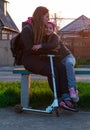 Mother and daughter on a bench in the park. Child riding on kickscooter. Blonde cute girl on scooter spring city street