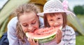 Mother and daughter eating watermelon near a tent in meadow or park. Happy family on picnic at camping Royalty Free Stock Photo