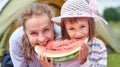 Mother and daughter eating watermelon near a tent in meadow or park. Happy family on picnic at camping Royalty Free Stock Photo