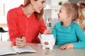 Mother and daughter putting coin into piggy bank at table indoors. Royalty Free Stock Photo