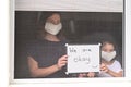 Mother and daughter in self isolation looking outside home window holding a sign reading: We are okay Royalty Free Stock Photo