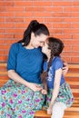 Mother and daughter in similar clothes hug and sit on the bench in front of red brick wall