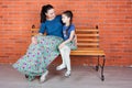 Mother and daughter in similar clothes sit on the bench in front of red brick wall.