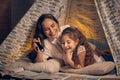 Mother and daughter are sitting in a teepee tent, reading stories with the flashlight. Happy family.