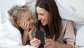 Mother daughter using smartphone laughing lying in bed under blanket