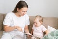 Mother explaining things while blond baby girl looking at book. Sitting on sofa in living room Royalty Free Stock Photo