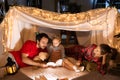 Mother, father and daughter in pajamas lying inside self-made hut, tent in room in the evening and reading book. Lovely Royalty Free Stock Photo