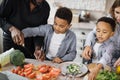 Mother and father teaching lovely kids to cook chopping fresh vegetables with knife.