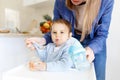 Mother feed little toddler baby boy with full mouth baby food, from plastic spoon in high chair view from back