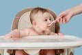 Mother feeding toddler baby with spoon on high chair for feeding children, blue studio background. Child at the age of six months Royalty Free Stock Photo