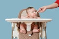 Mother feeding toddler baby with spoon on high chair for feeding children, blue studio background. Child, six months old, eats Royalty Free Stock Photo