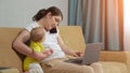 Mother freelancer works on laptop talks on phone holding kid Royalty Free Stock Photo