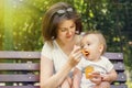 Mother giving her infant child complementary feeding pumpkin puree in sunny day outdoor. Both mom and kid sit on bench in park