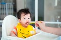 Mother hand holding small spoon Feeding Baby,toddler is messy wearing yellow bib apron and sitting In High Chair. Royalty Free Stock Photo