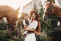 A mother in a hat with her son in her arms stands next to two beautiful horses in nature. a family with a child is photographed