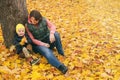 Mother and her little child boy sitting near huge maple tree in autumn park and looking on falling leaves. Fall nature banner with Royalty Free Stock Photo