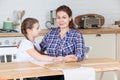 Mother and her ten years old daughter sitting together at table in kitchen, woman looking at camera, girl embracing her mom Royalty Free Stock Photo