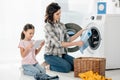 mother holding clothes near in washer wile daughter in pink t-shirt sitting with  digital tablet Royalty Free Stock Photo