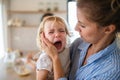 A mother holding a crying toddler daughter indoors in kitchen.